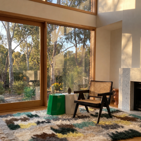 Modern interior with a chair, table, and large rug by windows overlooking trees.