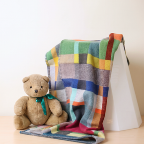 Multicolored checkered blanket draped over a white surface with a teddy bear sitting on it.