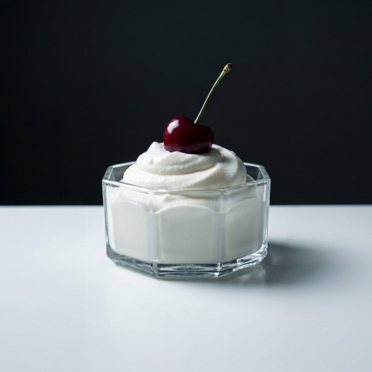 Octagonal clear bowl with cream and cherry on white table against a back backdrop