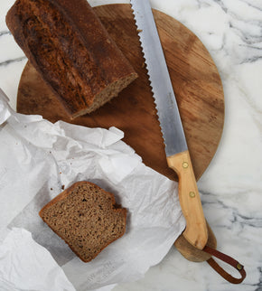 Pallares Solsona 25cm Box Wood Stainless Steel Bread Knife on rustic breadboard with dark brown bread loaf and slice