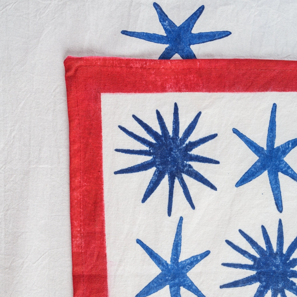 A close-up of a textile placemat showing a white fabric panel with blue starburst patterns and a red border, laid against a white background.