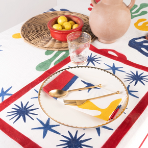 A styled table setting featuring a white tablecloth with a bold retro pattern of red borders, blue starbursts, and yellow abstract shapes. A matching red and white placemat with a gold-trimmed border is laid under a white plate with gold cutlery.