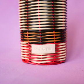 A close-up of a woven basket-style container or vase with alternating black, cream, orange, and red horizontal bands, featuring a cream label embossed with the brand name 'MAGEN' on a solid purple background.
