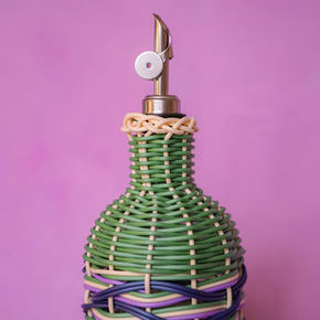 A decorative bottle with a green, purple, and beige woven rattan body, topped with a gold metal spout and a white circular stopper, set against a solid pink background.