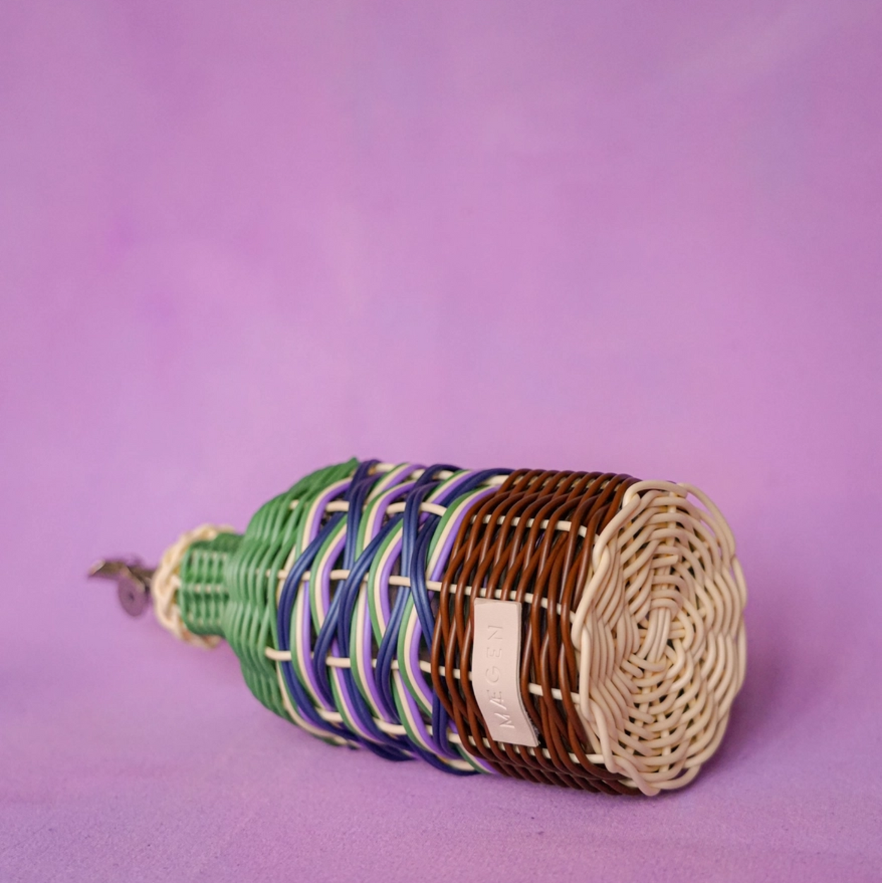 A handmade woven basket or scroll-like craft piece with green, blue, and brown woven bands, photographed on a solid purple background.