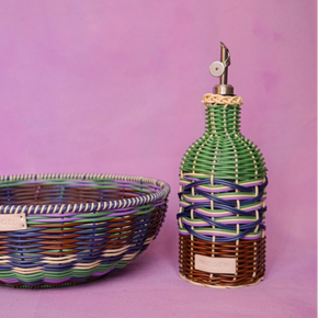 A hand-woven basket and a decorative bottle with a metal spout, both wrapped in multicolored striped raffia, set against a solid pink-purple background.
