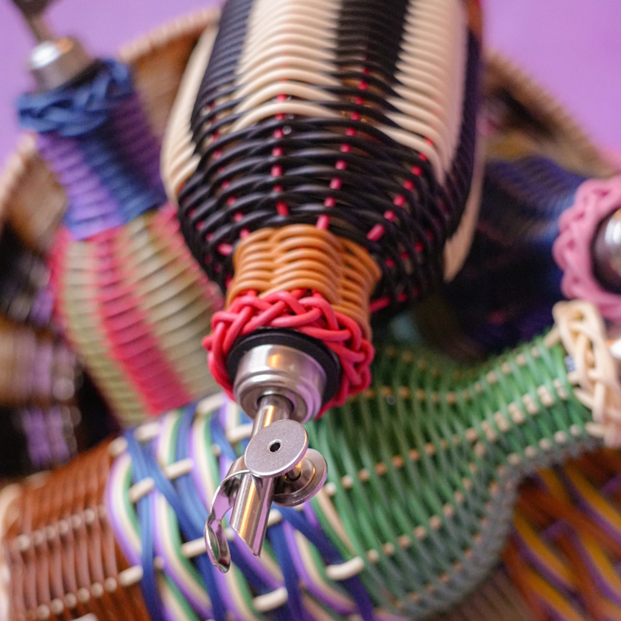 A close-up of a hand-woven basket filled with colorful woven cord oil bottles including black, white, red, blue, green, and purple tones, set against a solid purple background.