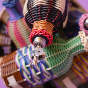 A close-up of a hand-woven basket filled with colorful braided rope or cord bundles, including black, white, red, blue, green, and purple tones, set against a solid purple background.