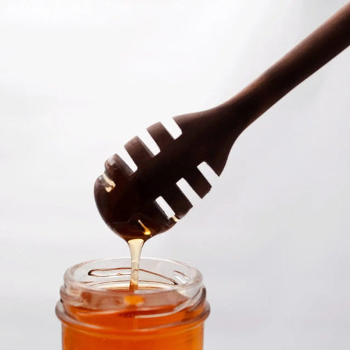 Honey drips from a wooden honey dipper into a glass jar on a white background