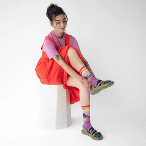Woman in red dress and green sandals wearing Bonne Maison Mauve Passage socks on white backdrop.