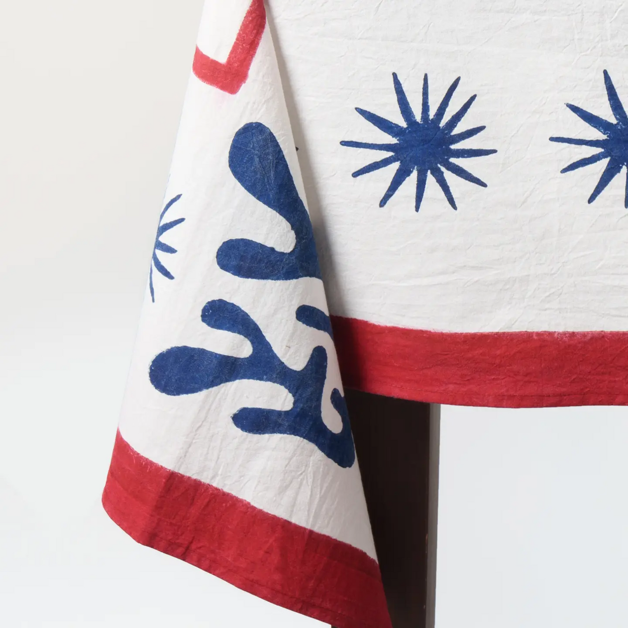 A close-up of a white textile, likely a tablecloth or napkin, featuring blue starburst patterns and red borders, draped over a dark stand against a white background.