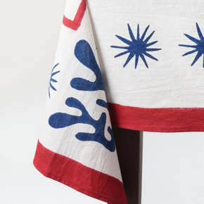 A close-up of a white textile, likely a tablecloth or napkin, featuring blue starburst patterns and red borders, draped over a dark stand against a white background.