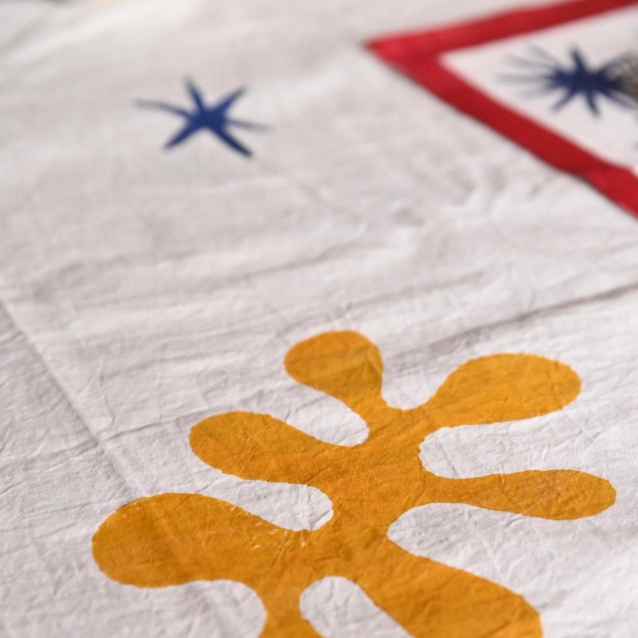 A close-up of a white fabric surface with a large mustard-yellow abstract flower-like motif in the foreground, plus partial blue and red starburst shapes in the background.
