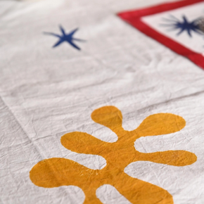 A close-up of a white fabric surface with a large mustard-yellow abstract flower-like motif in the foreground, plus partial blue and red starburst shapes in the background.
