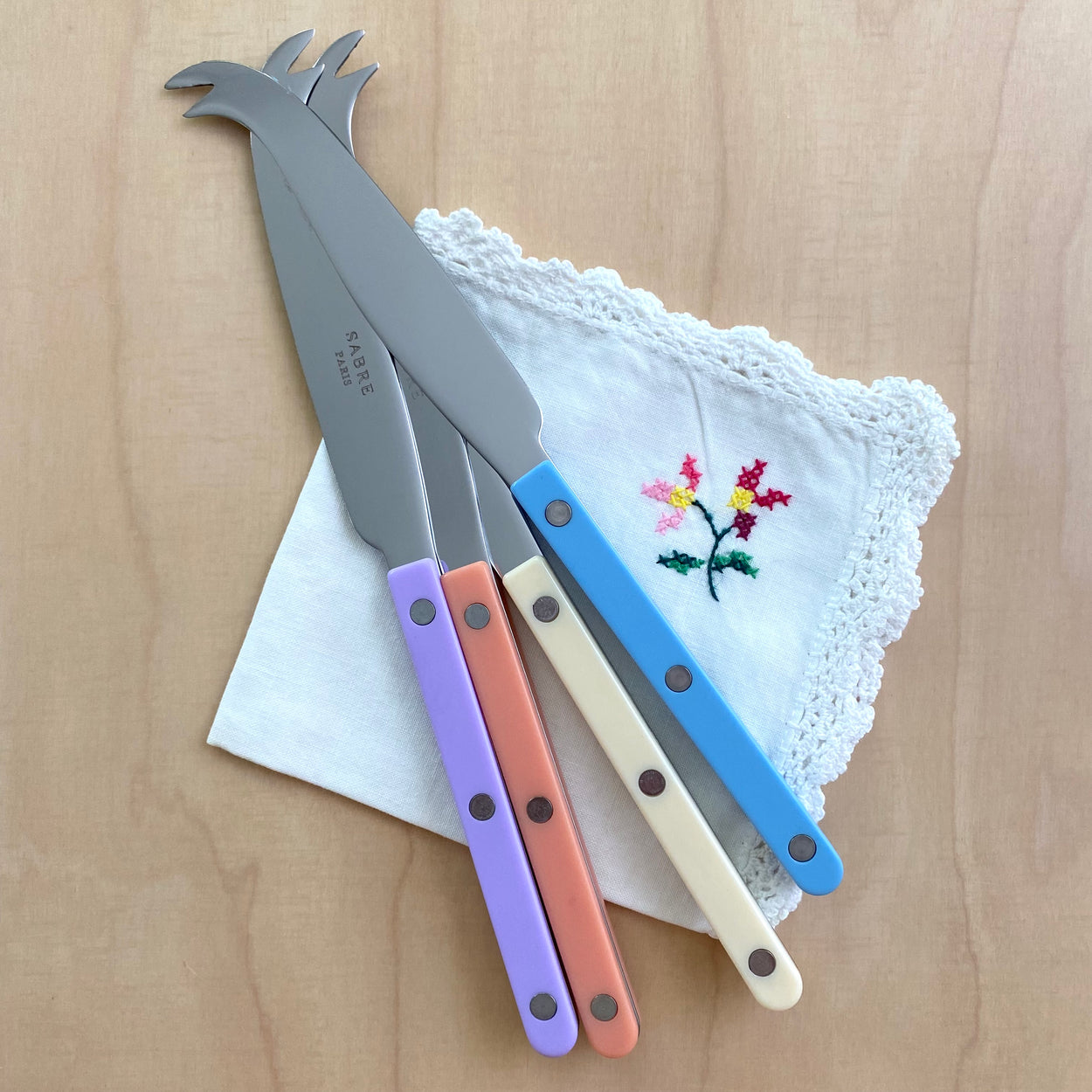 Sabre Bistrot Cheese Knife in Ivory, Pastel Blue, Lilac and Nude Pink on a hand cross stitched napkin.