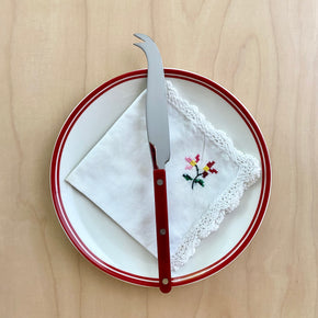 Sabre Bistrot Cheese Knife in Burgundy on a white plate with red trim and a hand cross stitched napkin