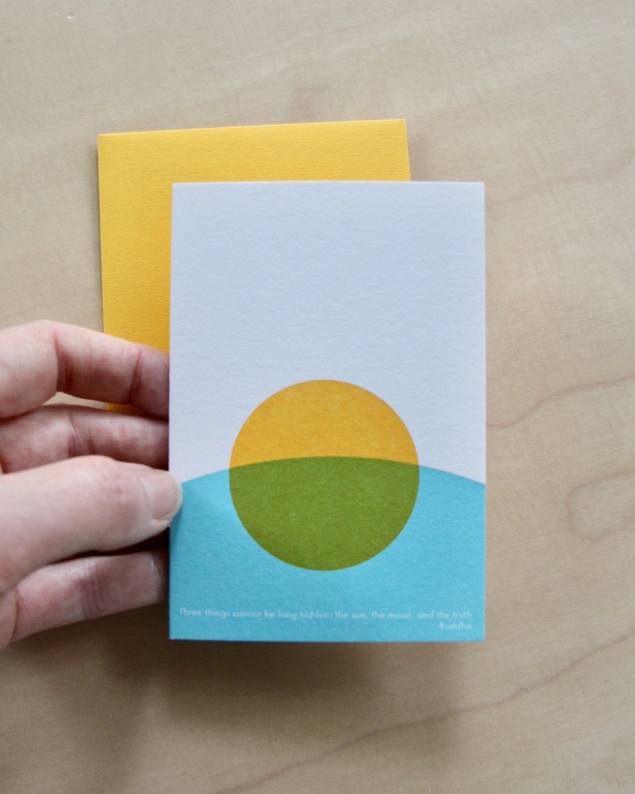 Hand holding a Hand printed greeting card with yellow sun and blue curve on a yellow envelope. reading "Three things cannot be long hidden, the sun, the moon and the truth" Buddha