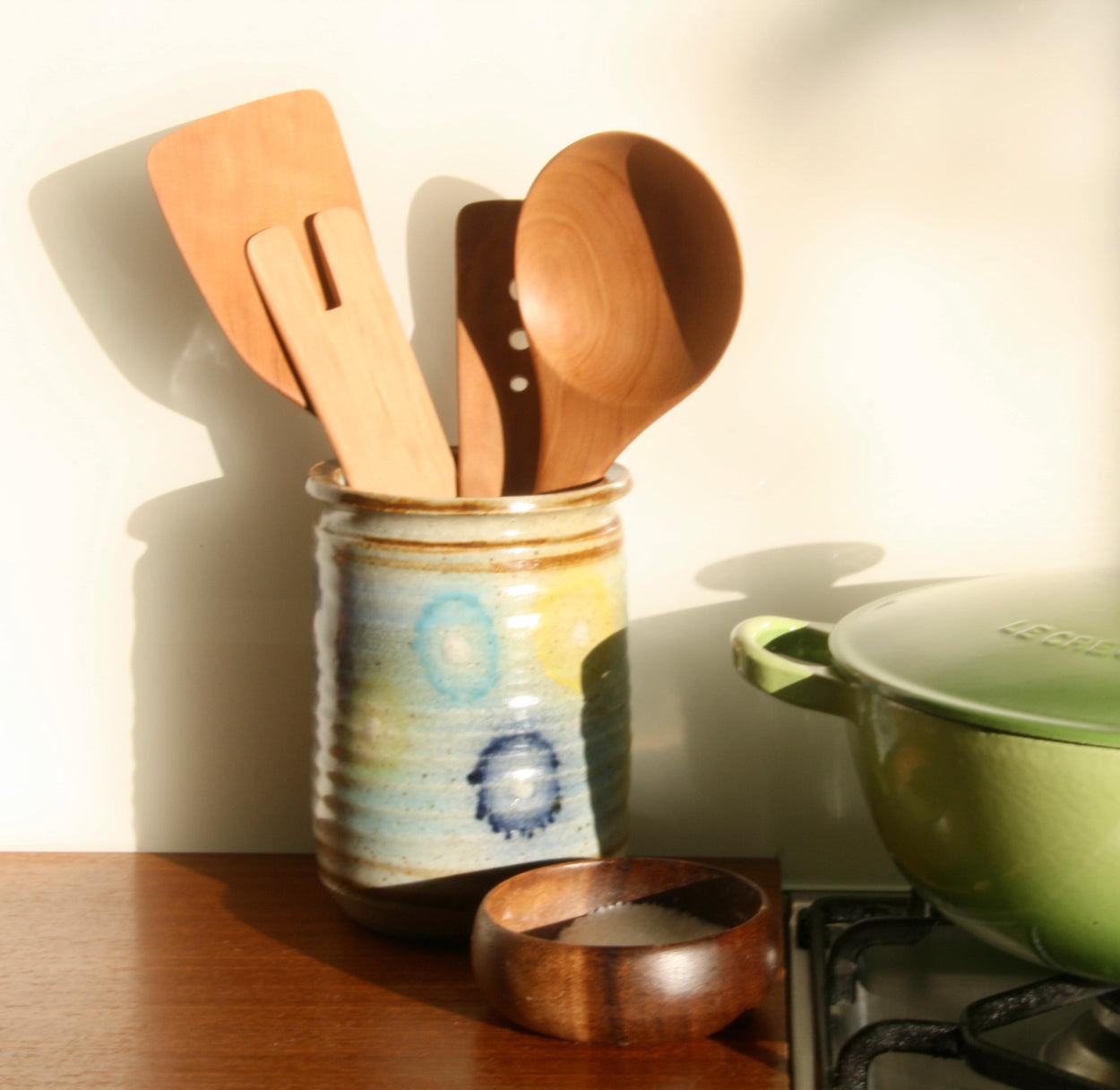 Handmade Pear Wood Soup Ladle with other Pear Wood kitchen utensil in ceramic kitchen utensil holder, next to green pot and salt dish.