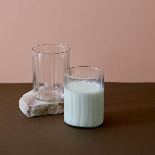 Hand crafted recycled glass tumblers x 2, one on a slab of marble, one 3/4 full of milk  against a pink and brown background.