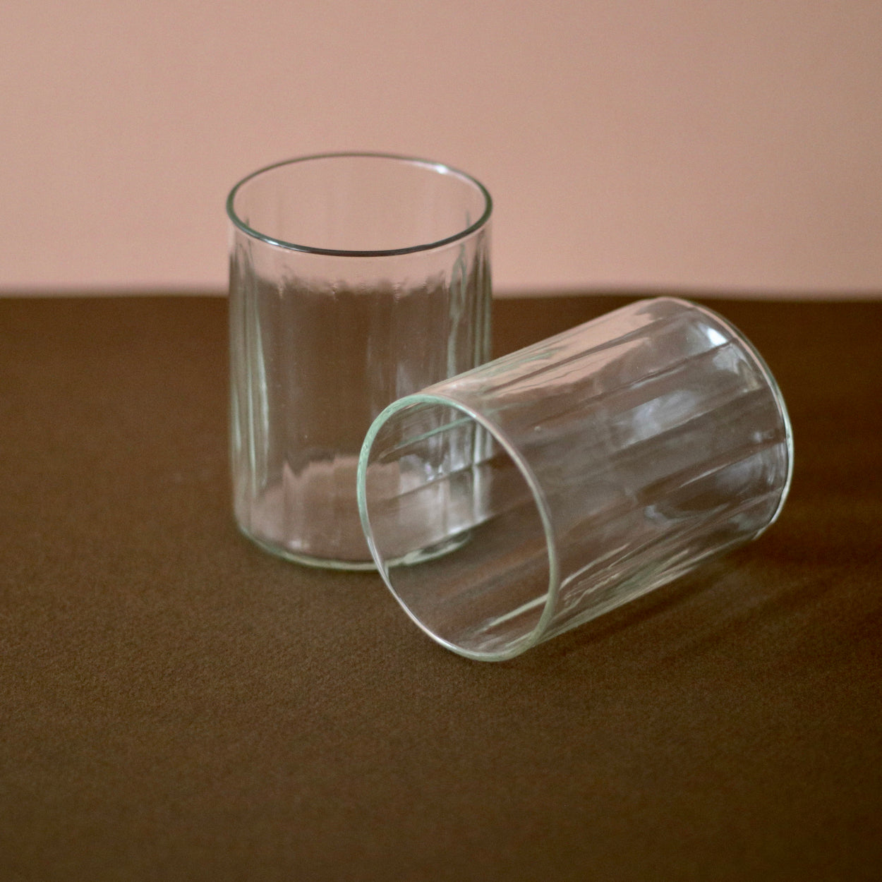 Hand crafted recycled glass tumblers x 2, one on its side, against a pink and brown background.