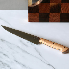 Pallares Solsona 17cm Aragon Carbon Steel Beech Wood Kitchen Knife on white marble bench with wood cutting board behind.