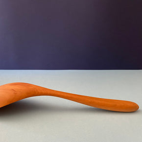 Handmade Pear Wood Soup Ladle lying against blue backdrop, face down.